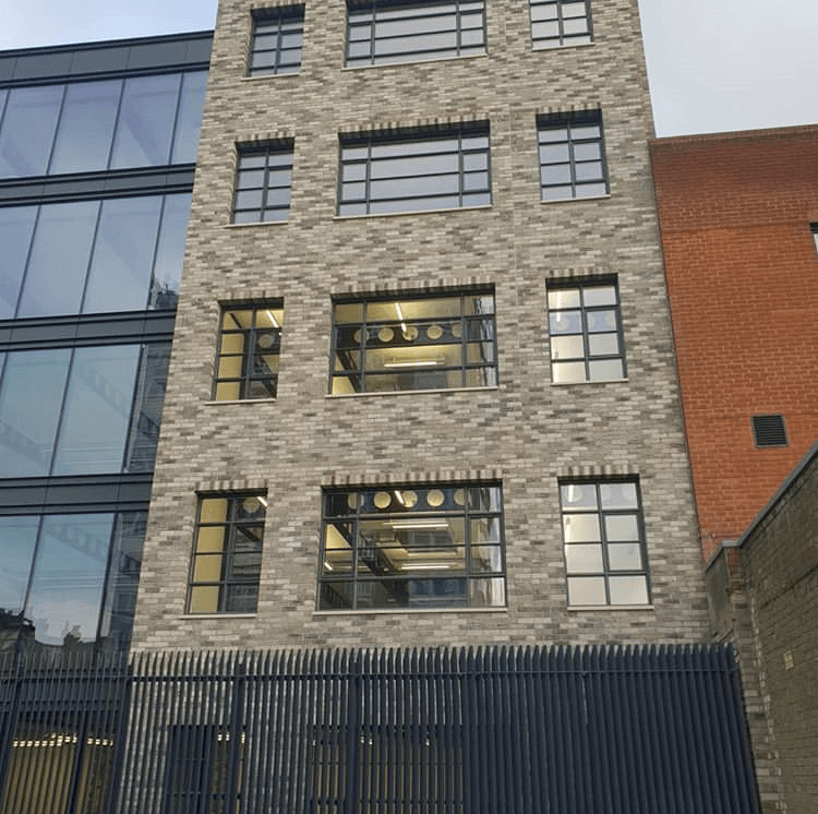 Modern building with a MCPV021 facade and large windows reflecting the cloudy sky, nestled between a glass building and an orange building.