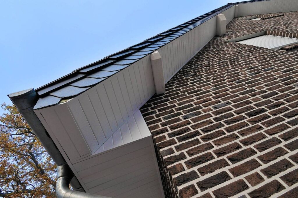 A low-angle view of a house facade featuring brick walls and a white soffit, complemented by black roof shingles from the MCP4033 product line. A metal gutter runs along the roof edge, with part of a tree visible on the left.