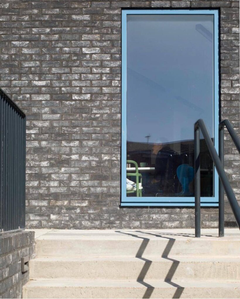 A large MCPV069 with a deep blue frame embedded in a dark brick wall, reflecting a clear sky, next to outdoor stairs with a black railing.