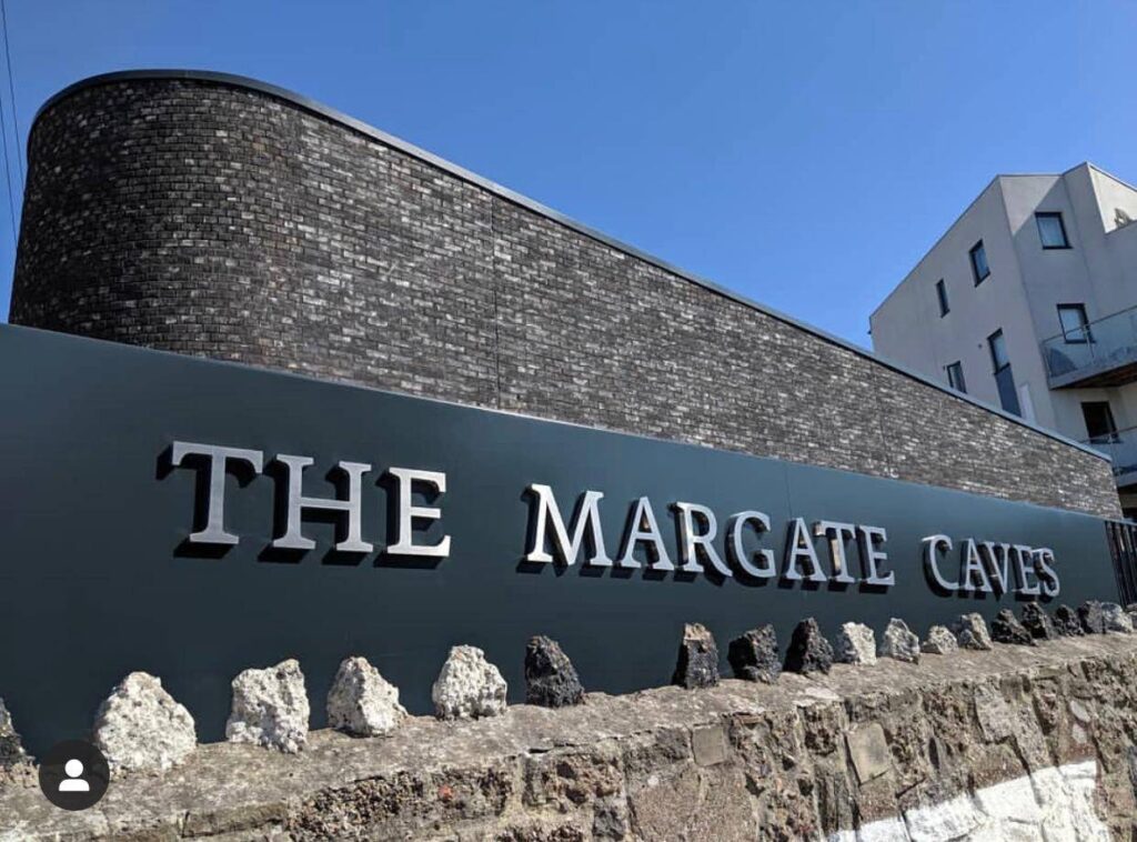Exterior of the MCPV069 with a curved stone wall and large lettered sign, under a clear blue sky.