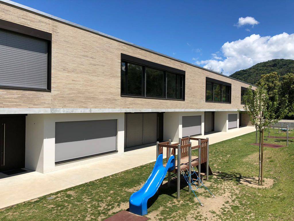 Modern building exterior with large windows and sliding doors, featuring a small playground with a MCPV352 slide in the foreground.