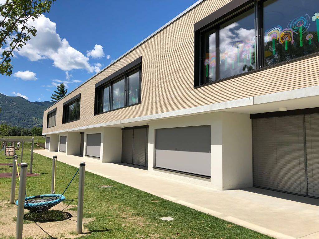 Modern two-story MCPV352 with large windows and beige walls, surrounded by a green lawn and mountains in the background.