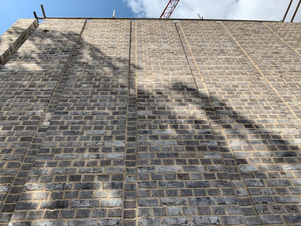 A large MCPV063 wall viewed from below with shadows cast by unseen objects, and a crane visible at the top edge against a blue sky.