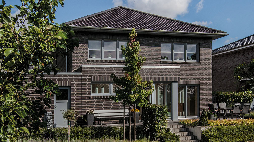A two-story brick house with a gabled roof and symmetrical windows, surrounded by lush greenery and a small patio area in the foreground of MCPH001.