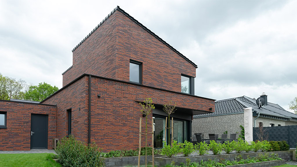 Modern two-story brick house (MCPH051) with large windows, surrounded by a landscaped garden and a cloudy sky overhead.