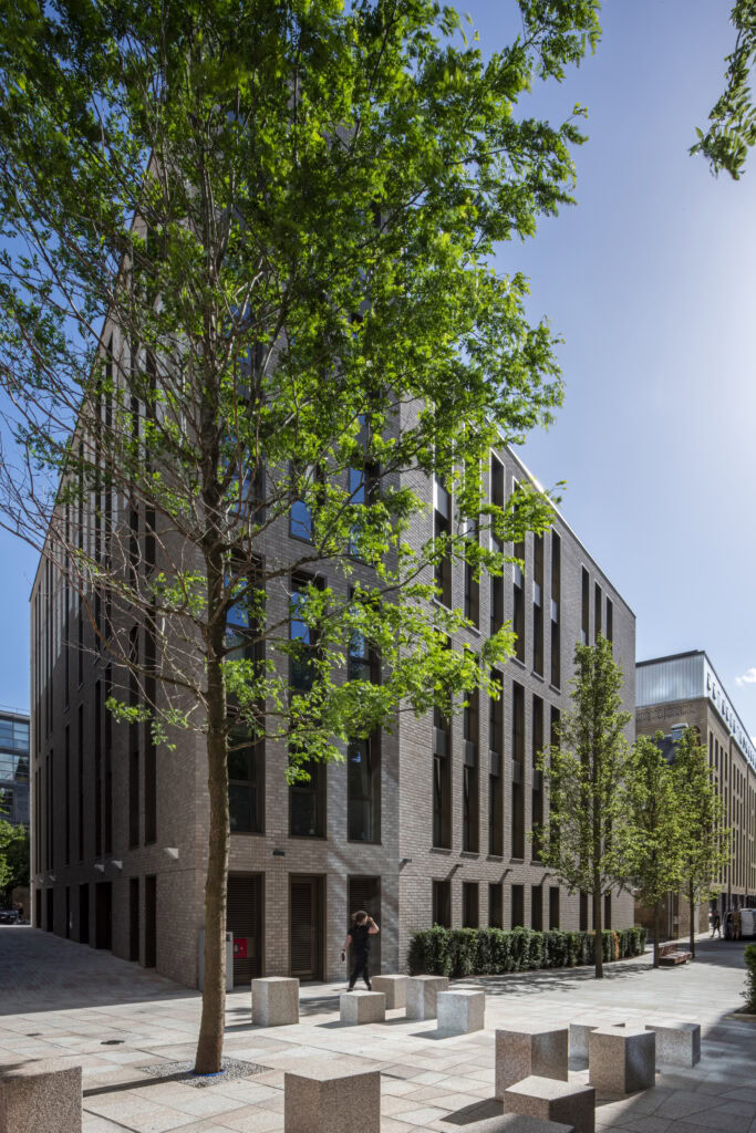 Modern urban office building with large windows and a brick facade, surrounded by young trees