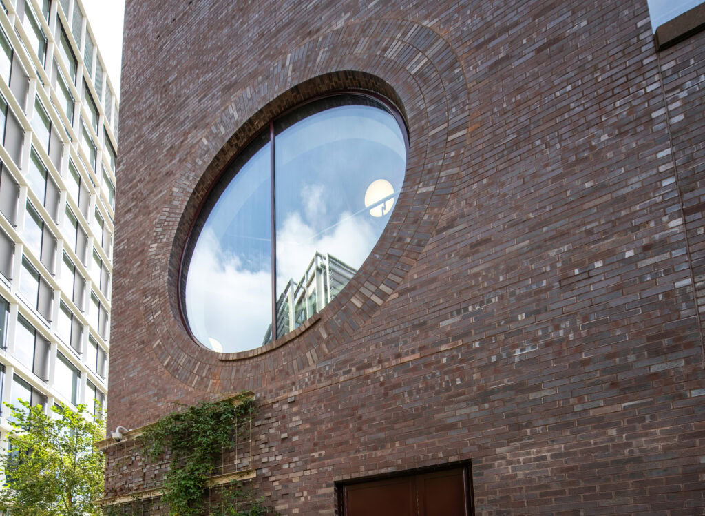 Large circular window in a brick building reflecting the sky and a nearby high-rise.