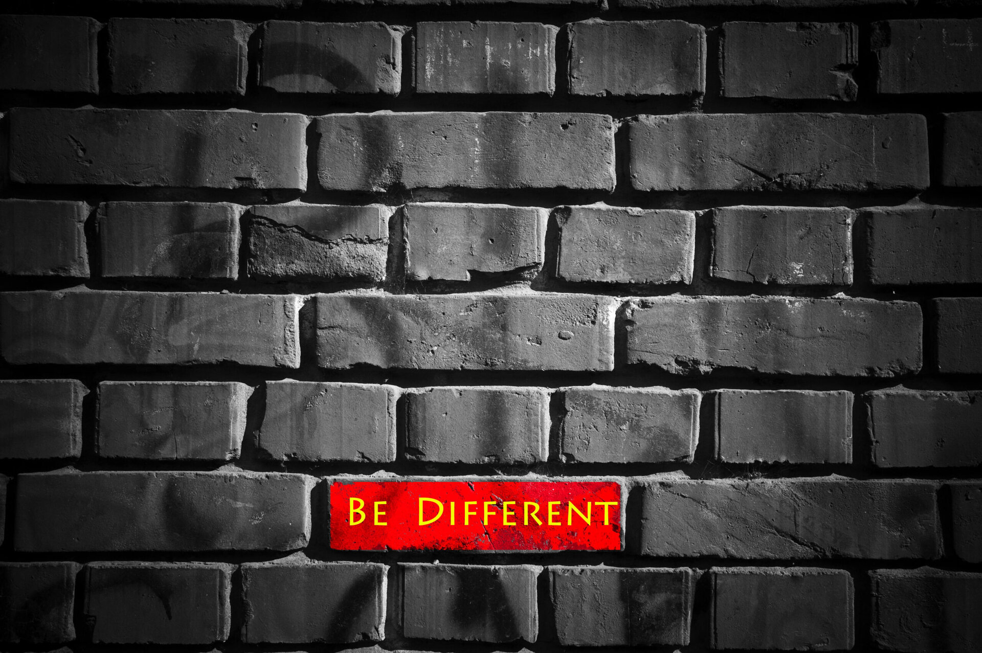 A black and white image of a brick wall with one brick in vibrant red, featuring the phrase "be different" in bold, white letters.