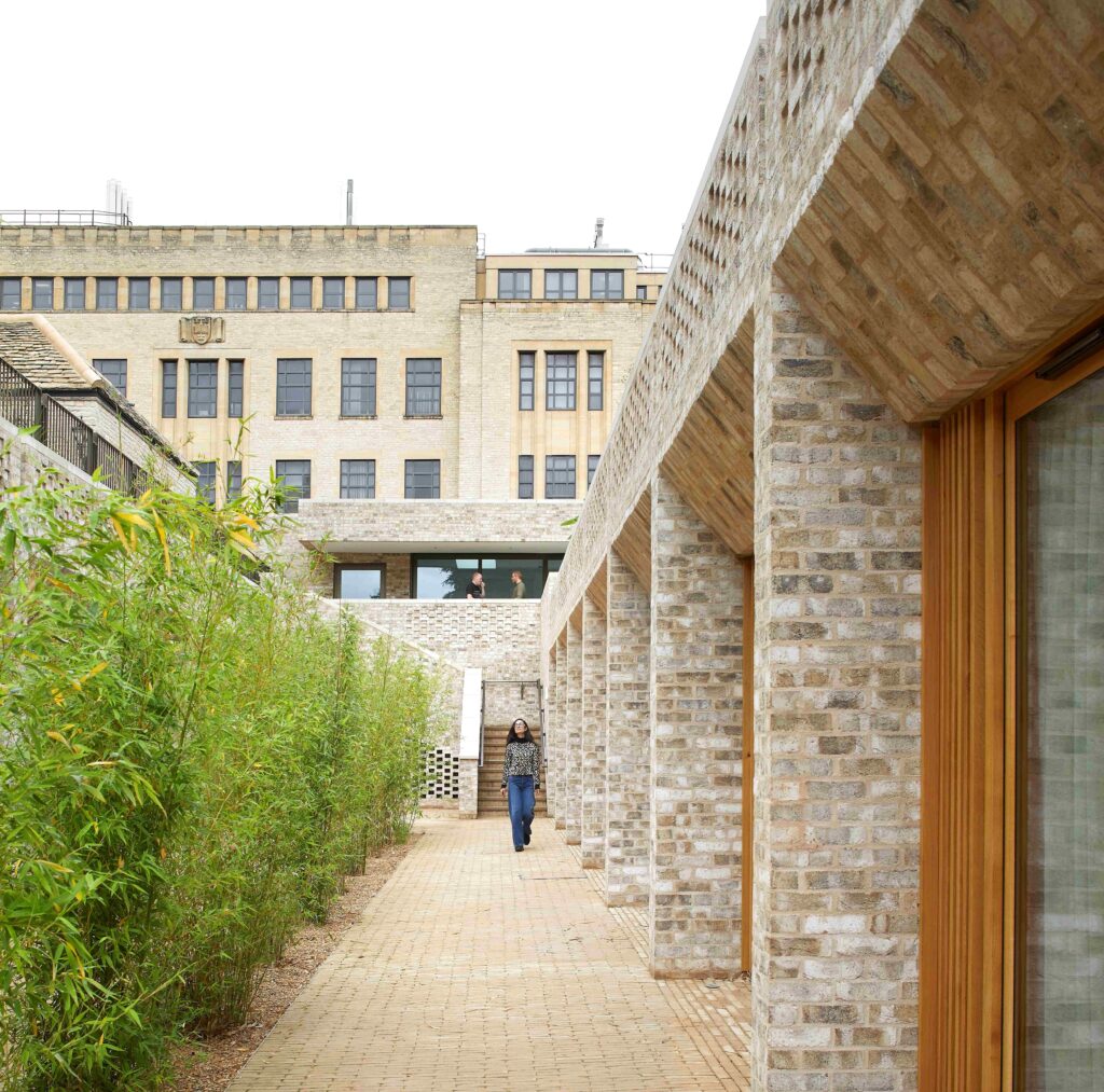 A woman walks down a narrow pathway adjacent to modern buildings featuring stone architecture and large windows.