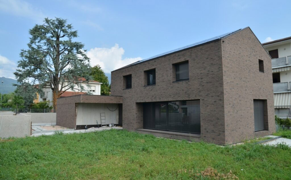 Modern brick house (MCPV038) with a flat roof and shuttered windows, standing in a grassy field with mountains in the background.